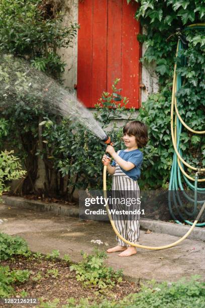 smiling boy watering plant with garden hose while standing on footpath at back yard - arroser photos et images de collection