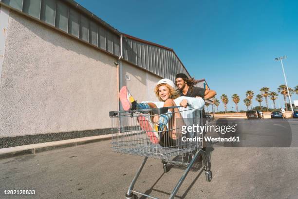 young man pushing girlfriend sitting in shopping cart outside supermarket - carro de corrida fotografías e imágenes de stock