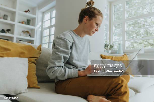 mid adult woman working on laptop while sitting on sofa at home - working from home stock-fotos und bilder