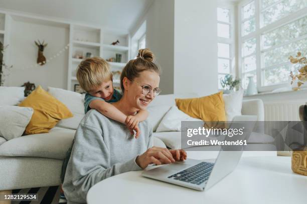 woman working on laptop while boy hugging her from behind at home - familie laptop foto e immagini stock