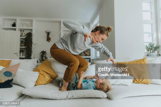 mother gesturing while standing on sofa by boy at home - family having fun stock-fotos und bilder