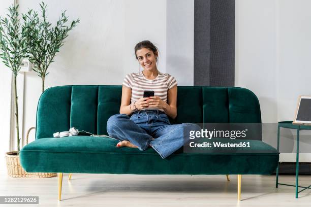 happy beautiful young businesswoman holding smart phone while sitting on sofa at small office - sofa stock-fotos und bilder