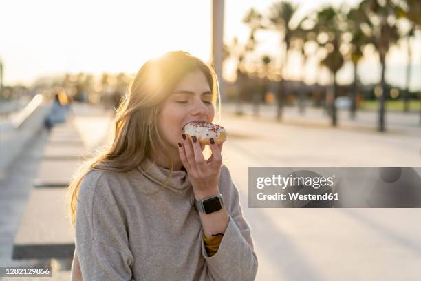 young woman enjoying fresh donut while sitting at promenade during sunset - eating donuts stockfoto's en -beelden