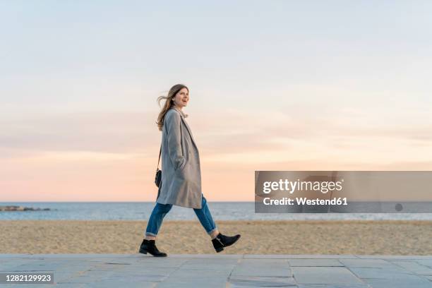 happy beautiful woman walking on promenade at beach while looking away against sky during sunset - woman walking full length stock pictures, royalty-free photos & images