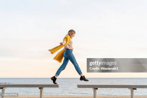 cheerful young woman jumping from bench to another at beach promenade against sky during sunset - jumping for joy stockfoto's en -beelden