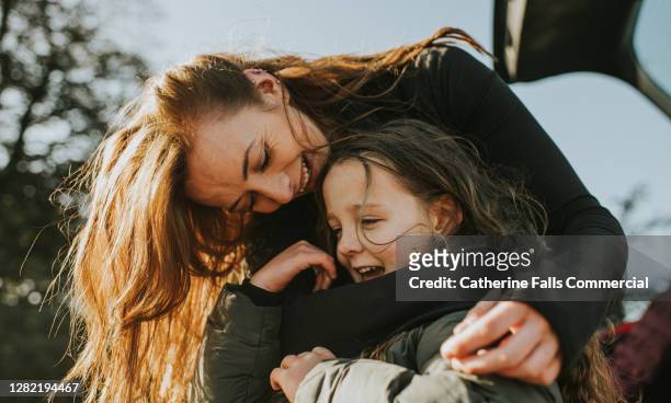 a mother bends down to embrace her daughter from behind - glücklichsein stock-fotos und bilder