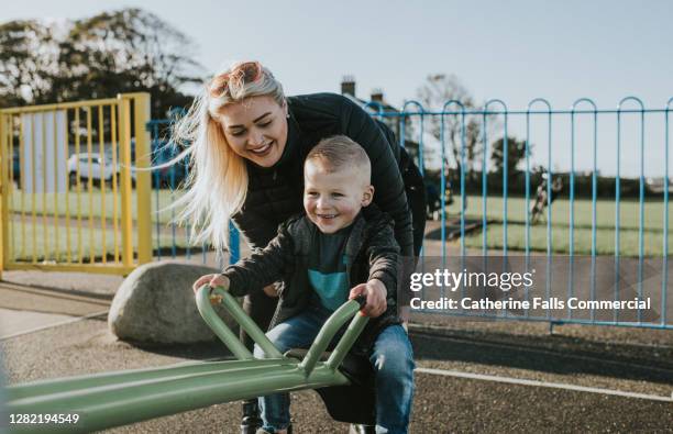 mother supports young son on a seesaw in a play park - kinderspielplatz stock-fotos und bilder