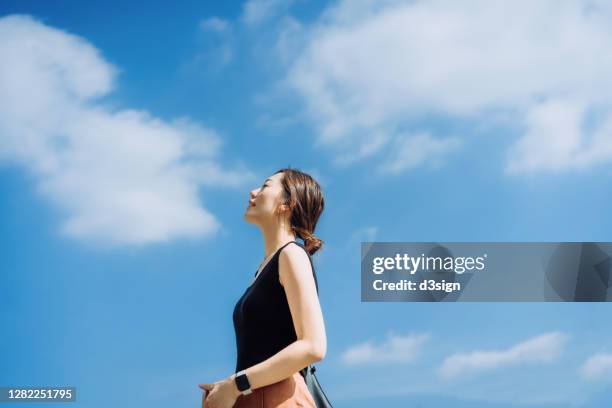 beautiful young asian woman relaxing in the nature, taking a deep breath enjoying some fresh air and gentle wind breeze with eyes closed, against clear blue sky on a sunny day - woman fresh air photos et images de collection