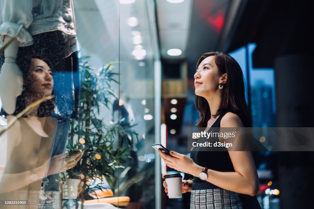 Cheerful woman holding a cup of coffee, checking her smartphone while standing outside a boutique looking at shop window in the evening in the city