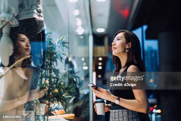 cheerful woman holding a cup of coffee, checking her smartphone while standing outside a boutique looking at shop window in the evening in the city - retail ストックフォトと画像