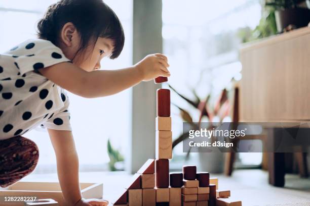 creative little asian girl crouching on the floor playing with wooden building blocks at home - montessori education stock-fotos und bilder
