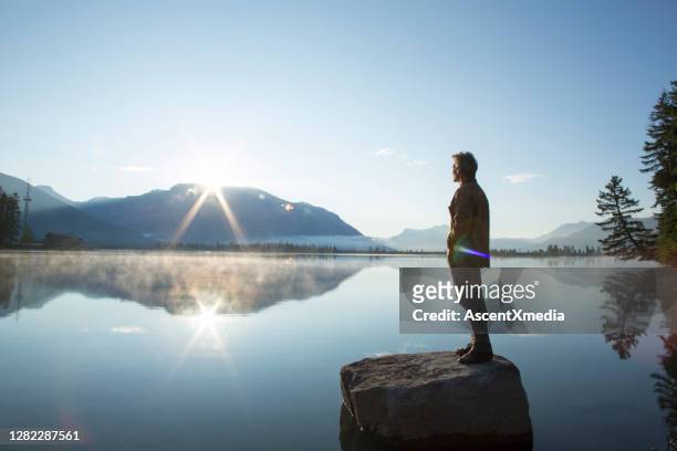 mature man stands on rock, in lake, and watches sunrise - tranquil scene stock pictures, royalty-free photos & images