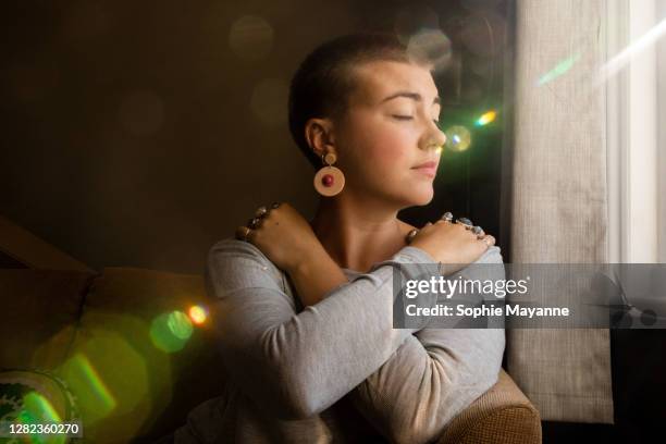 a young woman in her living room hugging herself in the sunlight - self-love stock pictures, royalty-free photos & images