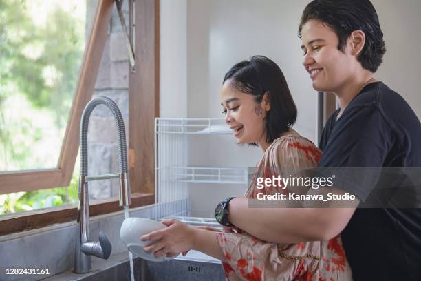 happy interracial lesbian couple cleaning dishes together - indonesia gay stock pictures, royalty-free photos & images