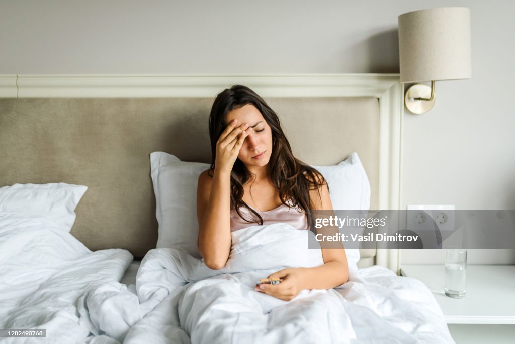 Young caucasian woman lying in bed, feeling unwell, with her hand on her head. Sickness / illness concept. Coronavirus / fever / headache concept. Home isolation.