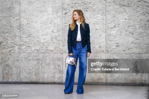 Guest wears earrings, a white ruffled mesh shirt, a dark navy blue jacket, a belt, blue denim flared jeans pants, a white bag, outside Chloe, during...