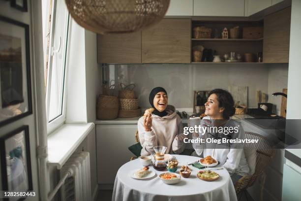 two happy multiracial girls having breakfast and drinking coffee at home together. multicultural friendship. one in hijab. - asian friends coffee stock-fotos und bilder