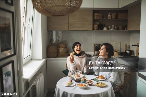 two happy multiracial girls having breakfast and drinking coffee at home together. multicultural friendship. one in hijab. - girl and coffee stockfoto's en -beelden