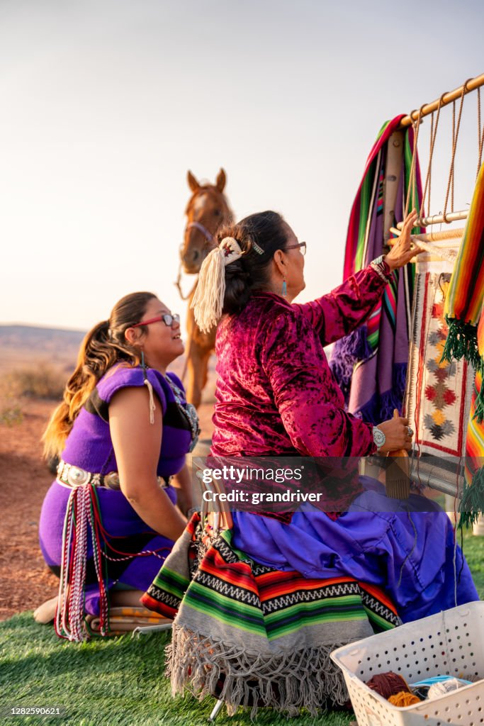 A Navajo Grandmother Teaching Her Granddaughter The Tradition Of Weaving On An Old Weavers Loom, wearing their traditional Navajo costume