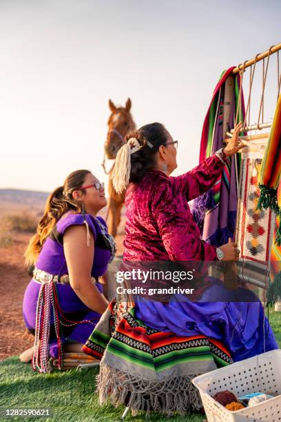 uma avó navajo ensinando a sua neta a tradição de tecer em um tear de tecelões antigos, vestindo sua tradicional fantasia navajo - tecendo - fotografias e filmes do acervo