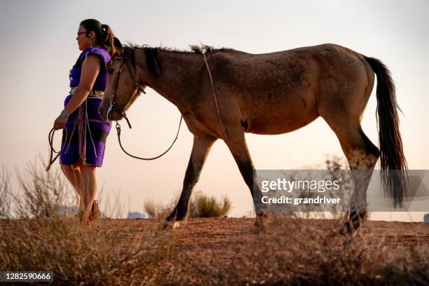 eine junge usnische amerikanerin trägt ihr natives kleid und türkis schmuck zu fuß mit ihrem pferd, das monument valley im hintergrund - lakota stock-fotos und bilder