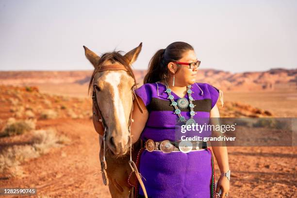 a young native american woman wearing her native dress and turquoise jewelry walking with her horse, the monument valley in the background - sioux native american tribe stock pictures, royalty-free photos & images