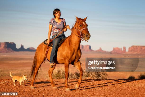 een jonge navajo die haar paard op een plateau boven haar huis met de vallei van het monument achter haar berijdt aangezien haar hond langs volgt - horse blanket stockfoto's en -beelden