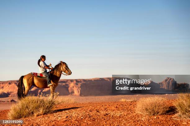 彼の馬を持つ若いナバホの少年は、ふざけて彼の家の上の高原に乗って、彼の後ろに記念碑の谷を持つ - ute ストックフォトと画像