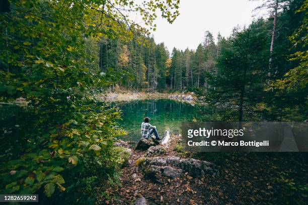 man sitting admiring an emerald lake in the forest - ambientazione tranquilla foto e immagini stock