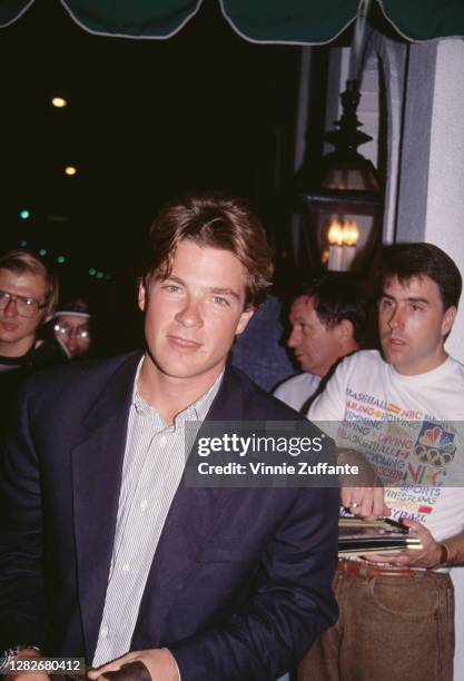 American actor Jason Bateman wearing a light grey shirt beneath a dark blue jacket, with a group of people in the background, one of whom wears an...