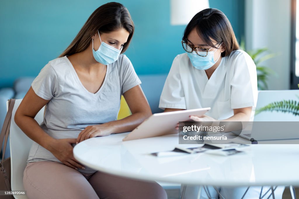 Young beautiful gynecologist wearing a hygienic face mask while showing to pregnant woman ultrasound scan baby with digital tablet in medical consultation.