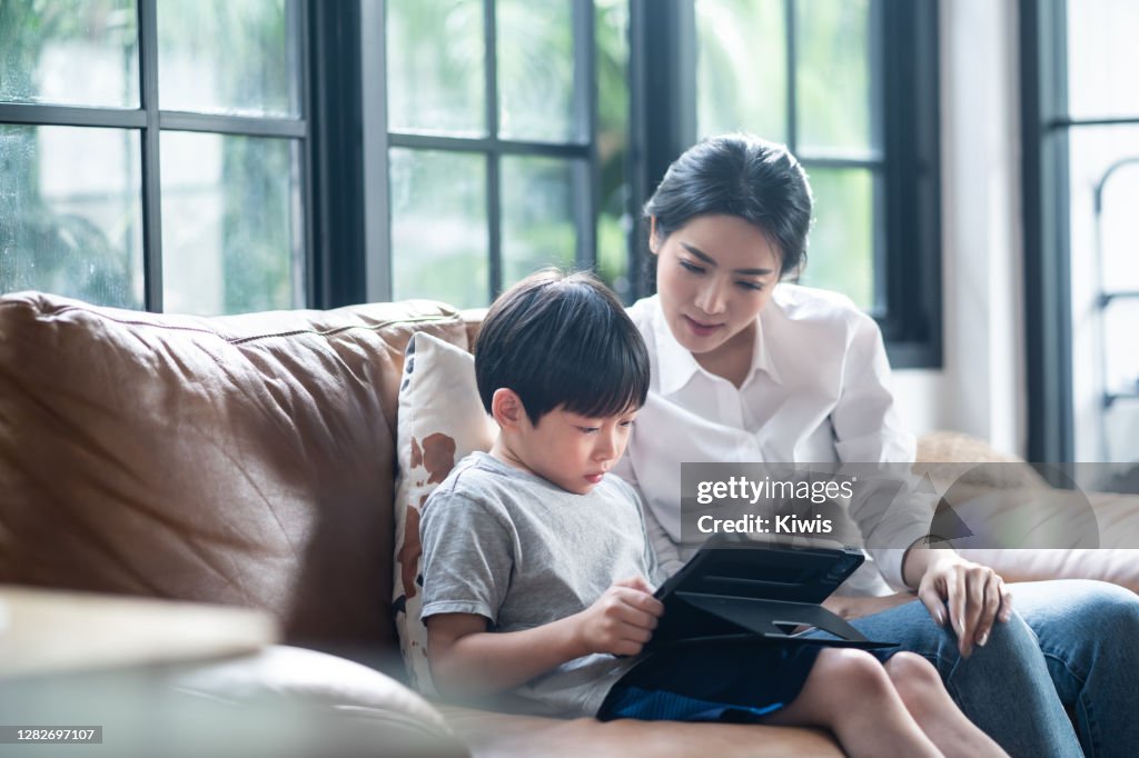 Asian mother teaching his young son for lesson on E-learning classes in tablet at home. The boy using tablet for learning. Remote education and social distancing to prevent covid infection.