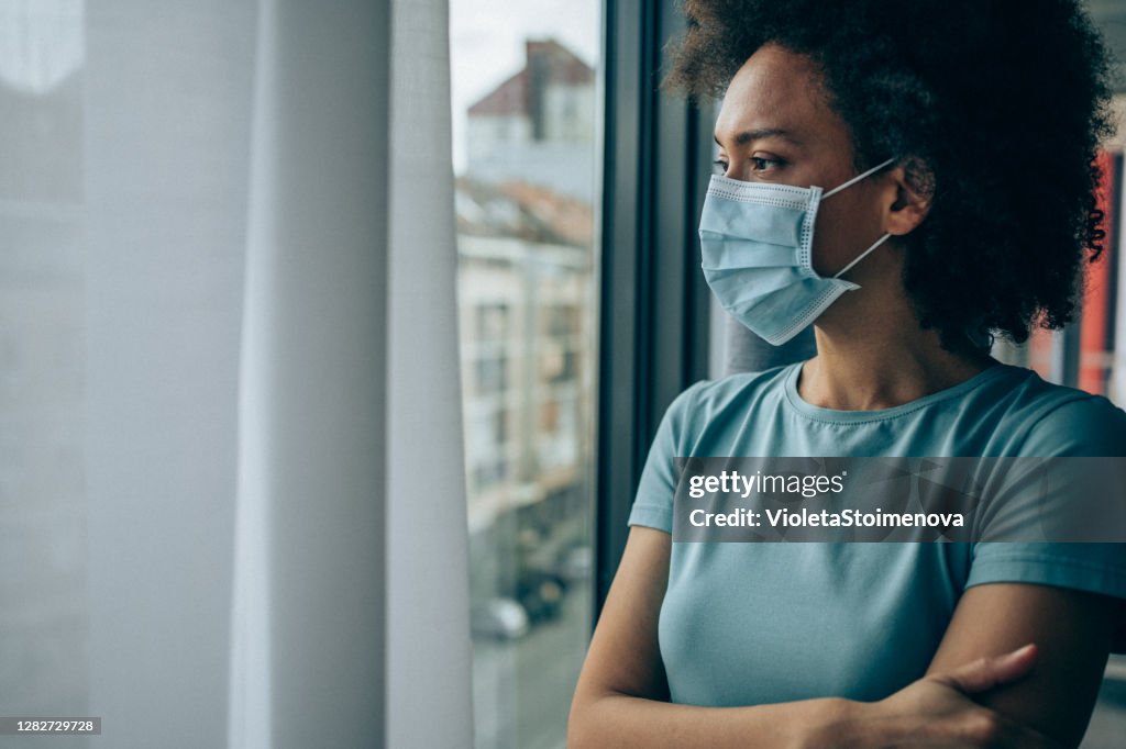 Worried young woman looking through window at home in quarantine.