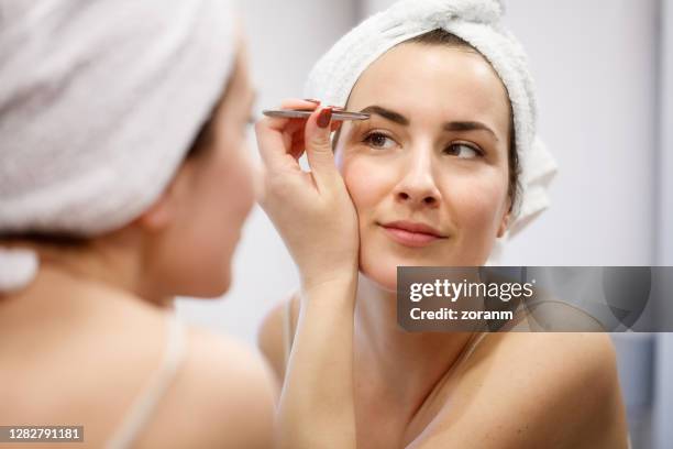 reflection of woman with towel on hair tweezing eyebrows - eyebrow tweezers imagens e fotografias de stock