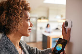 Mature Woman Using App On Phone To Control Digital Central Heating Thermostat At Home