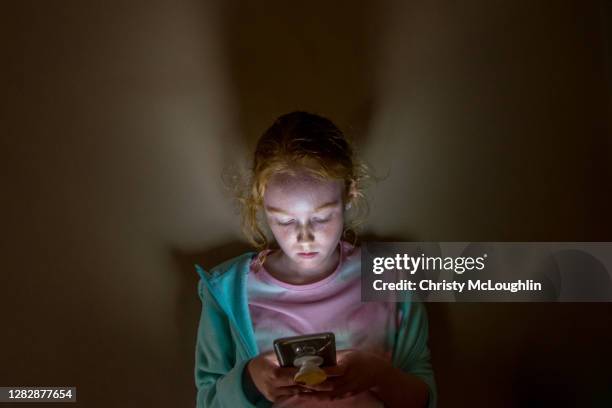 young girl looking into mobile phone screen, in dark room - bullying children stock pictures, royalty-free photos & images