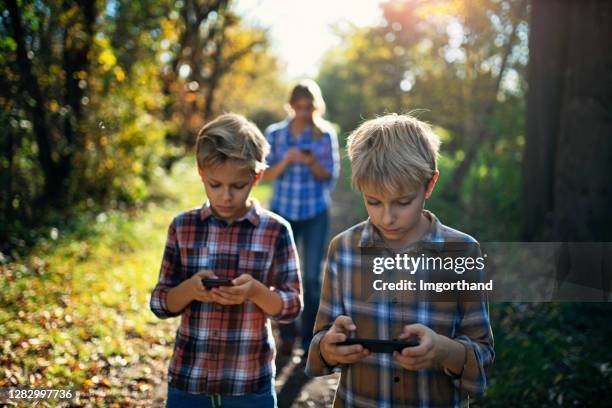 moeder en twee zonen die in stadspark met behulp van smartphones lopen - verkeerde-houding stockfoto's en -beelden
