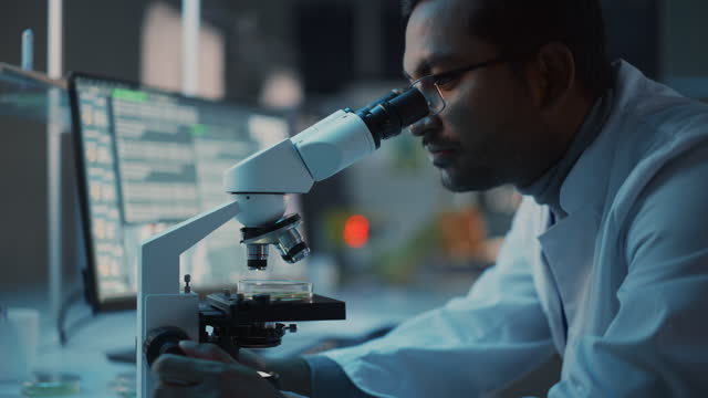 Medical Research Scientist Conducts DNA Experiments Under a Digital Microscope in a Biological Applied Science Laboratory. Multiethnic Lab Engineer in Glasses in White Coat Working on Vaccine.