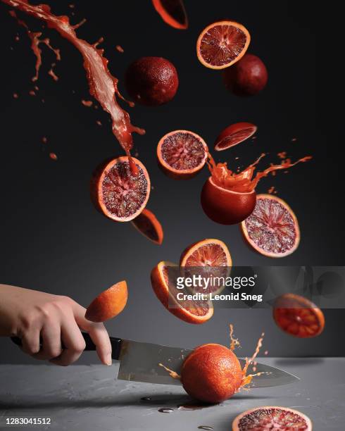 food photography of flying slices of red oranges with splashes of juice side view and a hand cutting fruit knife on a gray background close up - kitchen knife stockfoto's en -beelden