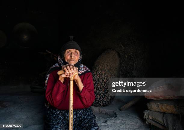 90-year-old senior woman wearing a headscarf sits in a dark room holding her cane - muslim woman darkness stock pictures, royalty-free photos & images