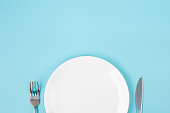 empty white ceramics plate with knife and fork on blue background. dining and kitchenware concept