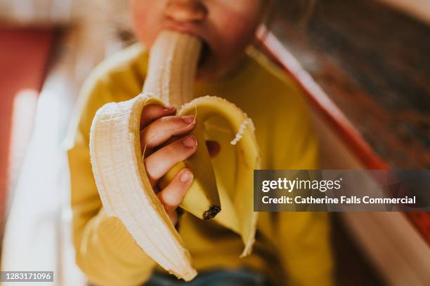 little girl in a yellow jumper eating a banana - banane essen stock-fotos und bilder