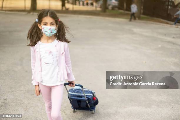 girl with a surgical mask walks looking at the camera dragging a wheeled backpack - südeuropa stock-fotos und bilder