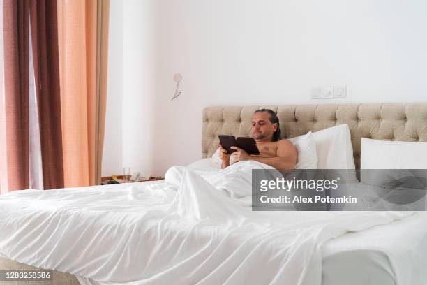 mature 50-years-old man lies in a bed and reading a book using e-reader. - 50 54 years imagens e fotografias de stock