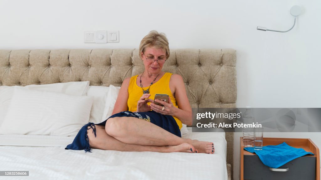 Mulher madura de 55 anos mandando sms com um smartphone quando ela estava sentada em uma cama em um quarto de hotel moderno e brilhante e limpo.