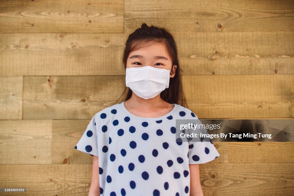 Lovely girl in protective face mask smiling joyfully at camera in front of a wooden wall