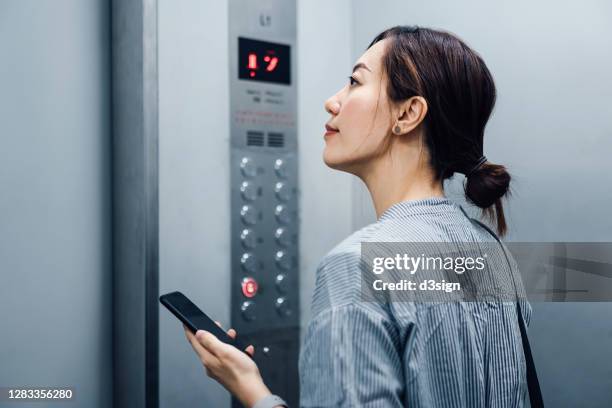 young asian woman using smartphone while riding in an elevator - ascensor interior fotografías e imágenes de stock