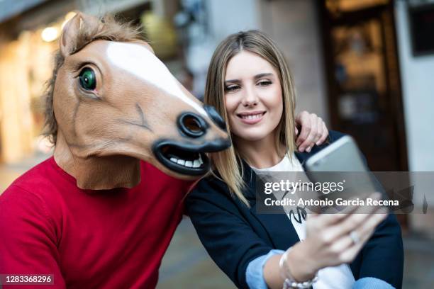 a strange date of a blonde woman with a man dressed as a horse, with a donkey mask outside a bar terrace taking selfie - ugly horses stock pictures, royalty-free photos & images