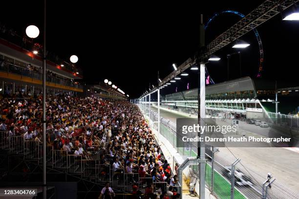 Thousands of Formula One fans and spectators in the main grandstand watch racing cars drive past on the start and finish straight opposite to the pit...