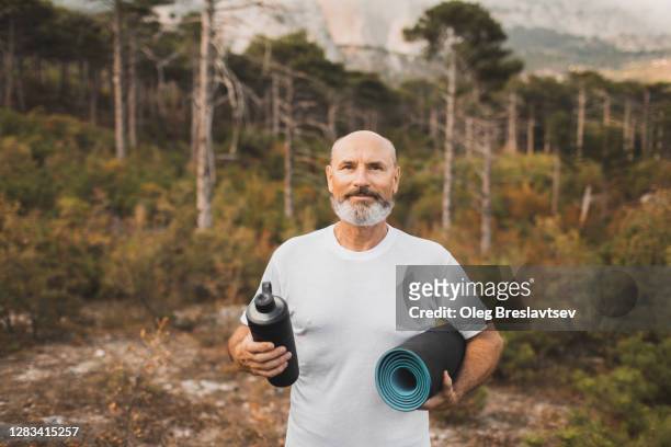 portrait of senior yoga coach with reusable water bottle and mat in white t-shirt outdoors in autumn forest. healthy lifestyle in old age - coach bus photos et images de collection